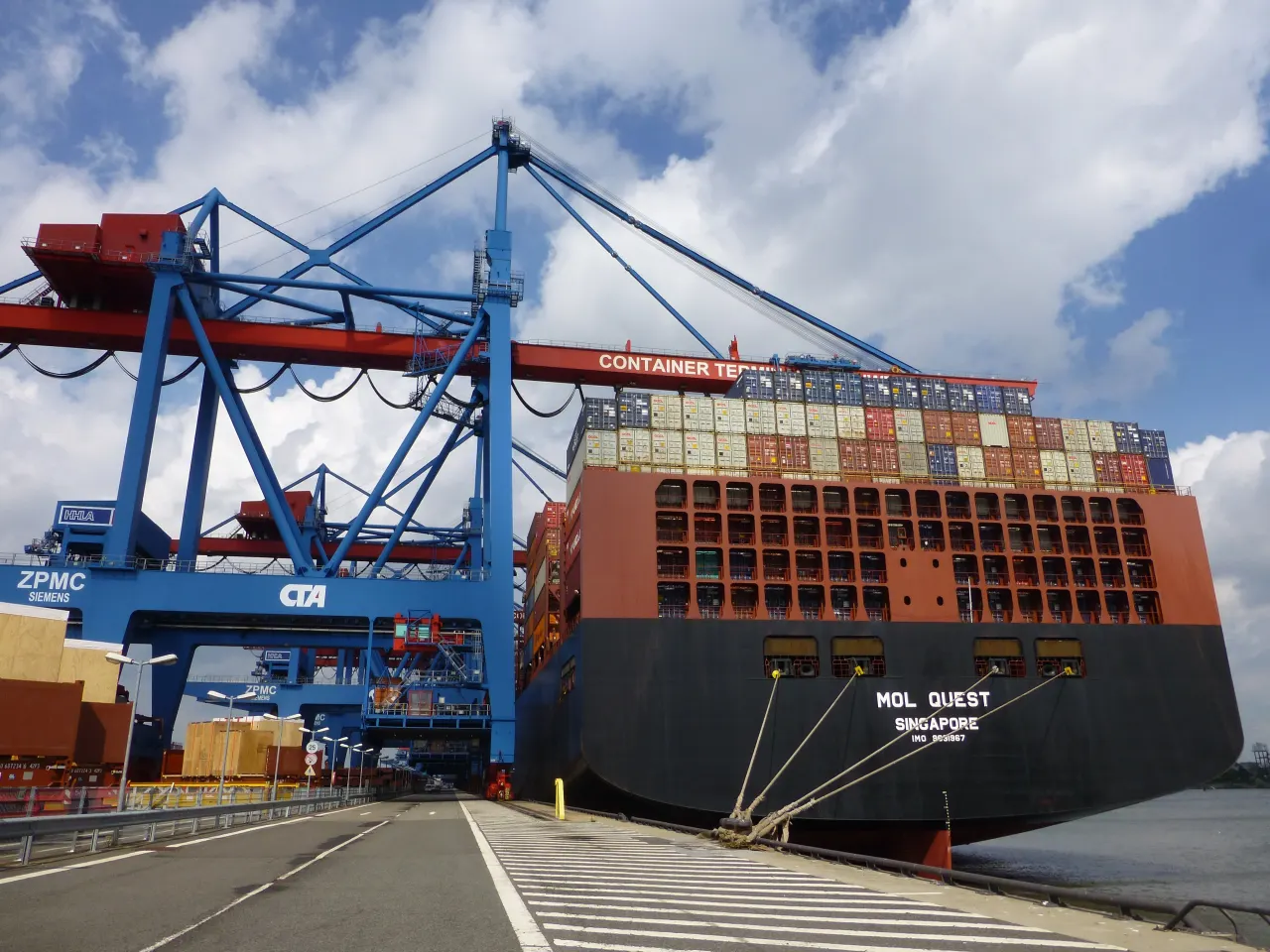 A loaded container ship moored under blue ship-to-shore cranes at a container terminal