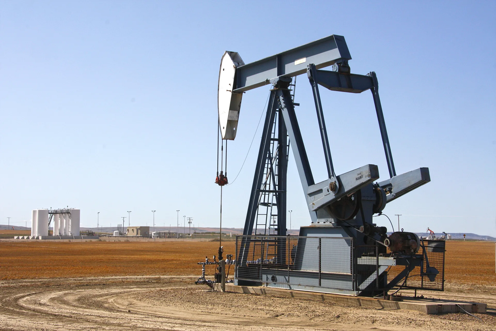 An oil pumpjack on a dry, harvested field, with storage tanks and more pumpjacks in the distance